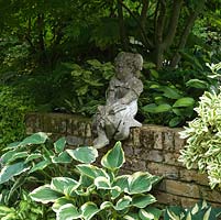 Small child statue sits on a wall in the shade of a large maple, above potted hostas.
