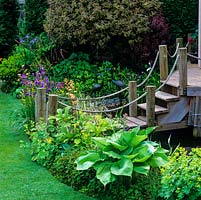 Raised wooden deck outside summerhouse is approached by steps with side posts linked by rose. Edged in Alchemilla mollis, hosta, Iris sibirica, mimulus and rodgersia.