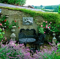 Sheltered by stone wall, sunken courtyard with wrought iron table, bench and chairs - wall with baskets and urns of geraniums. Edged in aromatic catmint. Behind, view of fields.