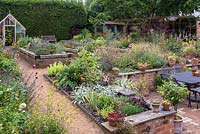 A split level, enclosed village garden with raised wooden beds planted largely with herbs, borders of late flowering summer perennials, a sunken terrace, pots of succulents and ornamental grasses, a wrought iron pergola and greenhouse.