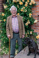 David Austin Roses. Portrait of Mr David Austin OBE VMH, accompanied by his Staffordshire bull terrier, Bertie. Behind, climbing on wall, Rosa 'Crown Princess Margareta'.