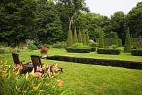 Orange Hemerocallis - Daylilies and pair of teak wood lounge chairs on manicured green grass lawn with Italian style mirror pool and clipped Thuja - Cedar trees and hedges in private backyard formal garden in summer