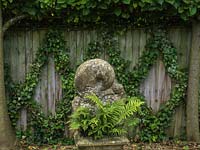 In shady corner, stone sculpture on plinth planted with ferns. Behind, a dull fence is cheered with a latticework of ivy.