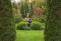 Manicured green grass lawn with water fountain, sculpture and crimson red Hydrangea paniculata shrubs framed by Thuja occidentalis 'Smaragd' - Cedar trees in private backyard garden in autumn
