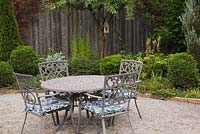 Brown cast iron metal round table and chairs on gravel stone patio and paving stone edged boders with globe shaped Thuja - Cedar shrubs, Prunus - Cherry tree with birdhouse in private backyard garden in autumn