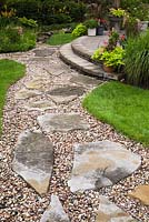 Flagstone and gravel path through manicured green grass lawn leading to raised brown paving stone patio with planters in residential backyard garden in summer