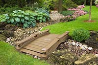 Manicured green grass lawn with brown painted wooden footbridge over dry stream and borders with Hosta plants and pink Impatiens flowers in residential backyard garden in summer