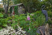 Heron sculpture in border with White Astilbe arendsii 'Weisse Gloria', Ligularia japonica, pink Digitalis flowers and old stone garden storage shed covered with climbing Menispermum canadense in private backyard garden in summer
