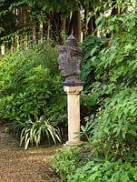 In shade of avenue of firs, stone head is raised on a plinth and set between borders of rhododendron, phormium, hardy geranium.