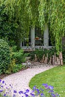 View over blue hardy geranium and curving stone path, to walkway crafted from lengths of birch by Claire Knights, sculptor. Beneath willow, shady bed of hardy geranium, tiarella and ferns