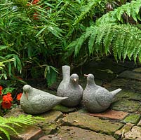 Three ceramic bird sculptures are tucked away on brick patio beneath ferns.