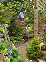 Collection of hostas and maples in raised beds. Salvaged street signs, an old station clock and chimney pot. Lantern Tree - Crinodendron hookerianum