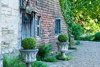 Grade II listed Jacobean manor house with fish scale tiles and front door framed with clipped box in stone urns amongst self seeded ferns and Alchemilla mollis. King John's Nursery, Etchingham, East Sussex, UK