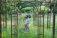 Stone putto framed by an ornate metal arbour  surrounded by meadow full of long grass, buttercups, cow parsley and species roses. King John's Nursery, Etchingham, East Sussex, UK