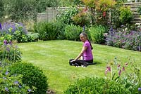 Nicola Stocken relaxes on the 8-week-old lawn in her newly rebuilt riverside garden, four months after it was devastated by February 2014's floods. Lawn enclosed in borders of Geranium Roxanne, box balls, Erysimum Bowles Mauve, delphiniums, Nepeta Walkers' Low, Sedum spectabile, Rosa Harlow Carr, Nandina domestica, Geranium psilostemon.