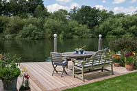 Riverside deck with dining table and chairs, is edged in pots of lillies and herbs, and beds of oriental lilies, daylilies, Verbena bonariensis, annual poppies, agapanthus, dogwood and kniphofia.