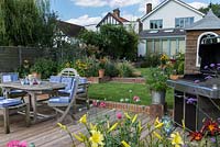 Riverside deck with barbecue, dining table and chairs, is edged in pots and bed of oriental lilies, daylilies, Verbena bonariensis and annual poppies. A step up leads to a lawn and raised bed. 