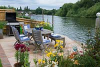 Riverside deck with barbecue, dining table and chairs, is edged in pots and beds of oriental lilies, daylilies, Verbena bonariensis, annual poppies, agapanthus, dogwood and kniphofia. Canoeists paddle by on the river.