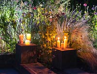 Candles and garden lighting illuminating a border at night, spotlighting Stipa tenuissima, annual poppy seedheads, Echium plantagineum, Nepeta Walker's Low and asiatic lilies.