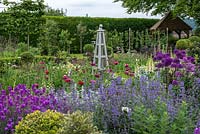 Wooden obelisk and vegetable garden beyond, seen over froth of Allium Purple Sensation, Erysimum  Bowles Mauve,  white valerian, aquilegia, foxgloves and catmint - Nepeta Walkers Low. Rising above are Ligustrum standards, privet cut into lollipop shapes.