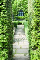 Path leads between yew hedges toward three grey painted chimney pots with a skirt of small leaved box, Buxus microphylla, in a steel container. 