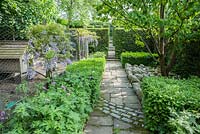 Path with curving cobble inset leads past a Davidia involucrata, the pocket handkerchief tree, mulched with large pebbles, and a chicken run decorated with standard wisterias underplanted with hardy geraniums. 