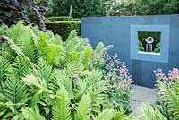 Ammonite sculpture by Darren Yeadon framed by a window in a grey wall trompe l'oeil - on one side of the flower garden, with lush ferns and pink astrantias in the foreground. Tony Ridler's garden, Swansea, Wales, UK