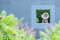 Ammonite sculpture by Darren Yeadon framed by a window in a grey wall on one side of the flower garden, with lush ferns and pink astrantias in the foreground. 