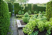 A path of paving slabs and stone setts, passes between lines of Christmas box, Sarcococca confusa, and raised beds containing box topiary shapes with architecture of tall yew hedges. 