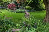 A stone sculpture of a bird on a plinth, surrounded with ferms, Alchemilla mollis self-seeding Aquilegias. Behind a small patio is surrounded with red Acers.