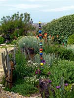 An exposed seaside gravel garden with raised island beds. Planting includes Helichrysum italicum, Briza maxima, Kniphofia 'Atlanta', hebe and a shelter belt hedge of Elaeagnus ebbingei. Decorative totem poles by local ceramicist Lynne Waylen.