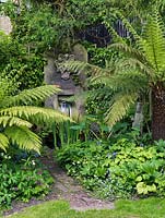 Water feature with waterfall gushing from huge stone into pool hidden in shady corner behind tree ferns