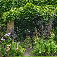 Pergola supported on brick columns, and covered in ivy, in bed of phlox, roses, sedum and lily, frames view of ivy covered plinth with pot of geranium.