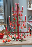 Wooden frame as a stylized Christmas tree, decorated with chilli bell peppers, rosemary, lighted red candles, ribbons and branches of olive, on countertop