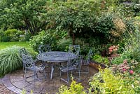 A circular stone patio with table and chairs. Planting in the surrounding raised bed includeds Sedum, Achillea, Stipa gigantea, Viburnum and a Sorbus tree.