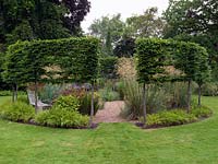 A round gravel garden cut out of a sunny lawn, encircled by a pleached hornbeam hedge. Summer.