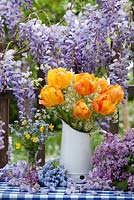 Outdoor spring display - tulips and cow parsley in jug, forget me nots and buttercups. Wisteria climbing against wooden fence.