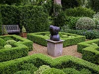 Knot garden, with four box topiary beds grouped around a central sculpture. Enclosed in hedges. White bench in conifer niche. Beyond, community garden.