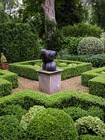 Knot garden, with four box topiary beds grouped around a central sculpture. Enclosed in hedges. Beyond, community garden.