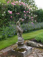 A stone statue stands in a corner of the herb garden. Behind, brick wall bearing climbing Rosa Aloha and Sombreuil - white above hardy geranium.