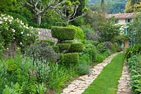 Grass pathway with stone edgings leads to villa through wide flower borders of front garden featuring a cloud pruned tree