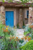 View of the blue door of the grass-roofed hut, with a handmade broom in front. Plants in the foreground include perennial wallflower Erysimum 'Apricot Twist', dyer's woad - Isatis tinctoria, Artemisia ludoviciana 'Valerie Finnis' and Salvia nemorosa 'Caradonna'. Sentebale - Hope in Vulnerability garden. RHS Chelsea Flower Show, 2015