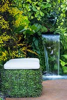 Green planted seat with white cushion sits in front of orchids and beside a clear cascade of water, pouring  from vertical planting or green wall growing  ferns. The Hidden Beauty of Kranji.  RHS Chelsea Flower Show, 2015