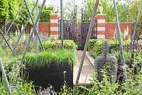 View through the garden from the sculpture of a figure through the metal poles and the rill to the gated entrance to the courtyard with the initials 'A' and 'W' on the gates The Living Legacy Garden by Darwin Property Management Investment. RHS Chelsea Flower Show, 2015