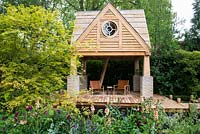 building, wooden deck patio with two chairs and bridge over pond surrounded by flowerbeds with Rosa 'Nuits de Young', Rosa 'Chianti', Salvia nemorosa 'Caradonna' and Verbascum 'Cotswold beauty' and Acer - The M and G Garden. RHS Chelsea Flower Show 2015