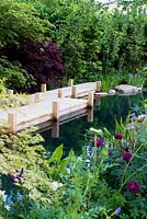The Retreat wooden board walk beside natural swimming pool with black water backed by hedge and cottage-style border in front. The M and G Garden 2015 -  RHS Chelsea Flower Show 2015