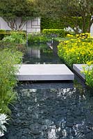 The Telegraph Garden, view of concrete block path and bridge over modern pond  with black slate bottom leading to concrete feature walls surrounded by Doronicum x excelsum 'Harpur Crewe' and Allium moly and Osmanthus x burkwoodii 