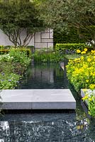 The Telegraph Garden, view of concrete block path and bridge over modern pond with black slate bottom leading to concrete feature walls surrounded by Doronicum x excelsum 'Harpur Crewe' and Allium moly and Osmanthus x burkwoodii 

