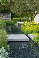 View along rill edged in beds of grasses, irises and perennials - golden doronicum and euphorbia on right. The Telegraph Garden. RHS Chelsea Flower Show, 2015
