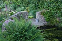 End of reservoir pond with stone wall, small jetty and watering-can. Fern in foreground, Zantedeschia aethiopica 'Crowborough'AGM - arum lilies behind wall